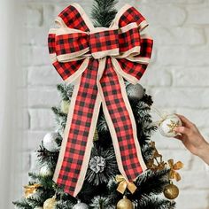 a christmas tree with a red and black plaid bow on it's top, next to a hand holding an ornament