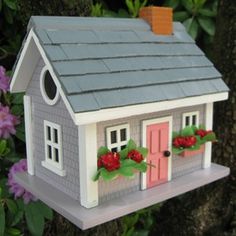 a birdhouse with flowers on the roof and windows is hanging from a tree in front of purple flowers