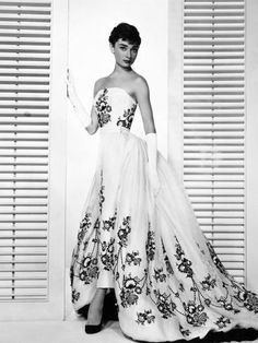 a black and white photo of a woman wearing a dress with flowers on it, in front of shutters