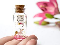 a hand holding a tiny bottle filled with pink and gold confetti next to flowers