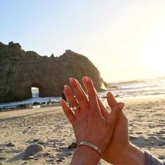 a person holding their hand up to the sky on a beach with an arch in the background