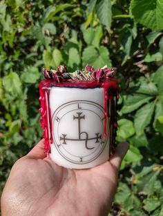 a hand holding a white and red ring with an anchor on it in front of green leaves