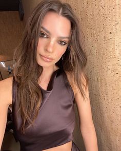 a woman with long hair wearing a brown top and posing for the camera in front of a wall