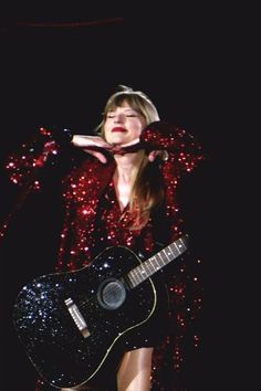 a woman in a red dress holding a black guitar and singing into a microphone on stage