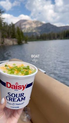 a person holding up a cup of soup in front of a boat on the water