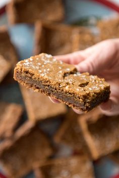 someone holding up a piece of brownie in front of a plate full of cookies