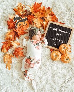 a baby laying on top of a white blanket next to pumpkins and a sign