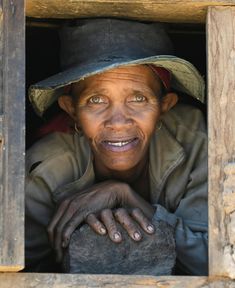 an old woman wearing a hat looking out from behind a wooden window with her hands on the ledge