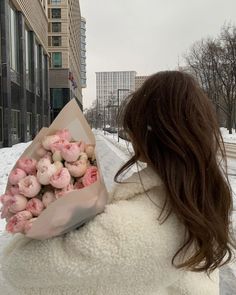 a woman holding a bouquet of pink flowers in the middle of snow covered street next to buildings