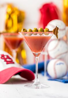 a pink martini with olives and garnish on the rim in front of baseball caps