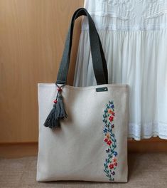 a white bag with embroidered flowers and tassels on the handle, sitting next to a curtain