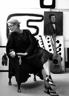 a woman sitting in a chair with her legs crossed and shoes on, next to paintings