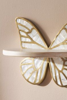 a white and gold butterfly shaped clock mounted to the side of a wall next to a wooden shelf