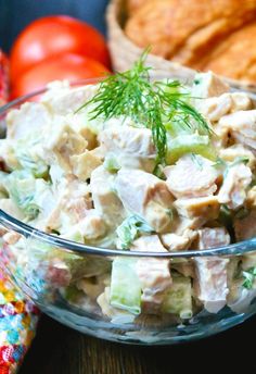 a glass bowl filled with chicken salad next to some bread and tomatoes on the side