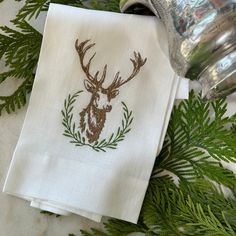 a white napkin with a deer head embroidered on it next to evergreen branches and a silver canister