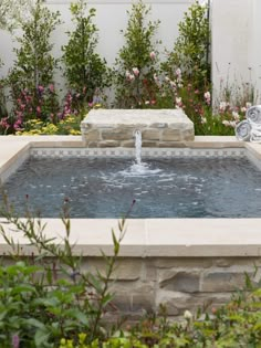 a small fountain in the middle of a garden with pink flowers and greenery around it