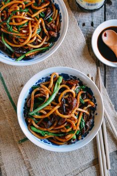 two bowls filled with noodles and vegetables next to chopsticks on a wooden table