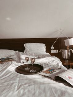 two glasses of wine sitting on top of a bed next to an open magazine and lamp