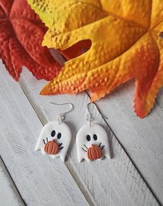 a pair of halloween ghost earrings sitting on top of a wooden table next to an orange leaf