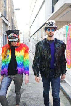 two people walking down the street wearing colorful clothing and feathers on their head, with one man in black jacket