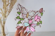 a hand holding a pink and green butterfly brooch in front of a vase with dried flowers