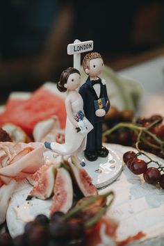 a cake with figurines on top of it and fruit in the background,