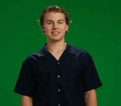 a young man standing in front of a green background wearing a black shirt and jeans