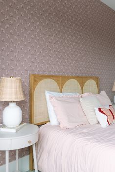 a bed with pink sheets and pillows next to a white night stand in front of a floral wallpaper