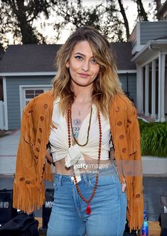 a woman standing in front of a house wearing jeans and a fringe jacket with beads