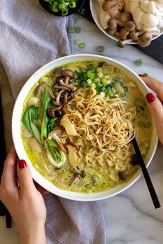 two hands holding a bowl of noodles with mushrooms and broccoli