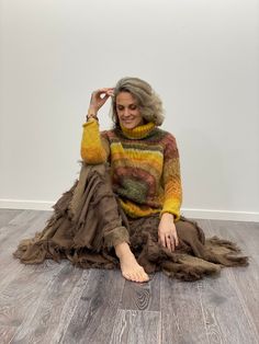 a woman sitting on the floor wearing a multicolored sweater and brown skirt with her hands in her hair