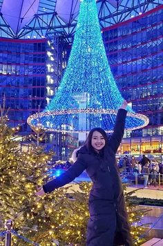 a woman standing in front of a christmas tree with lights on it and her arms up