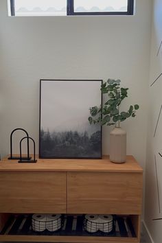 a wooden cabinet with two pictures on it and a plant next to it in front of a window