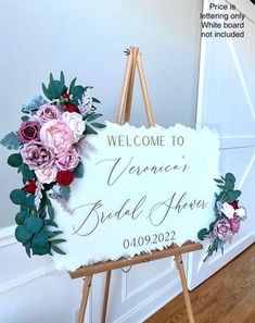 a welcome sign with flowers on it in front of a white wall and wooden easel