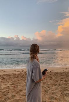 a woman standing on top of a beach next to the ocean holding a cell phone