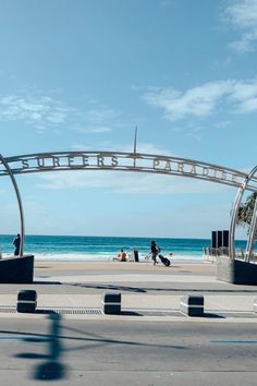 people are walking on the beach under an arched metal sign over looking the water and blue sky