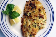 a white plate topped with fish next to a slice of lemon and green leafy garnish