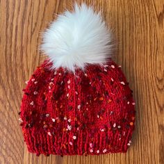 a red knitted hat with white fur pom - pom sitting on top of a wooden floor