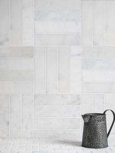 a black watering can sitting on top of a white tiled floor next to a wall