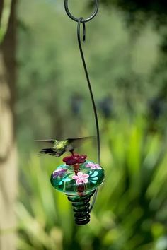 a hummingbird on a bird feeder with flowers in the foreground and a tree in the background