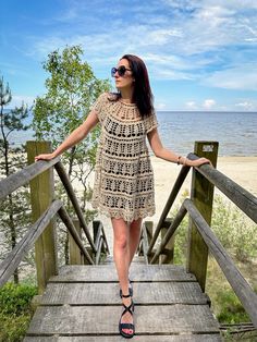 a woman is standing on a wooden bridge near the water and sand, wearing sunglasses