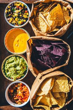 mexican food is served in paper bowls on a wooden tray, including tortilla chips and guacamole