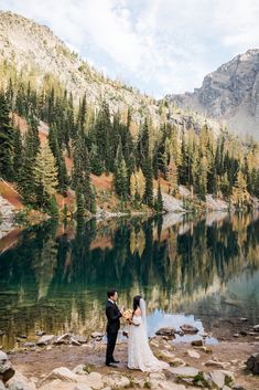 a bride and groom standing on the shore of a mountain lake