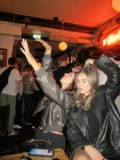 two women in leather jackets are dancing at a party with other people behind them and one woman is holding her arms up