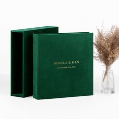 two green wedding guest books next to a vase with dried grass in it on a white background