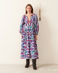 A woman is captured against a white wall wearing the Celeste Oasis Ikat Dress, a long, hand-printed cotton piece adorned in shades of blue, purple, and pink. The dress showcases a deep V-neck and puffy sleeves. She elegantly pairs it with black boots as a decorative tassel adorns the wall beside her. Cowry Shell, Ikat Dress, Gathered Neckline, Ikat Print, Printed Cotton Dress, Feather Light, Plunging Neckline, Xl Dress, Cotton Dress