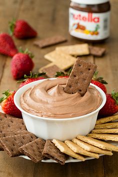 a white bowl filled with chocolate dip surrounded by crackers and strawberries on a wooden table