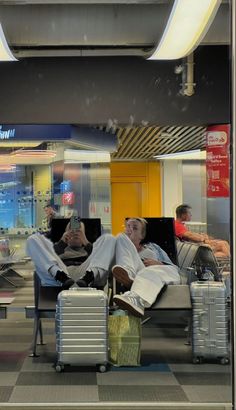 two men sitting on couches with luggage in front of them and one man holding a cell phone