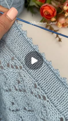 someone is knitting a blue sweater with a white button on the bottom and a pink rose in the background