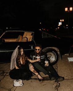 two women sitting next to each other on the ground near a parked car at night